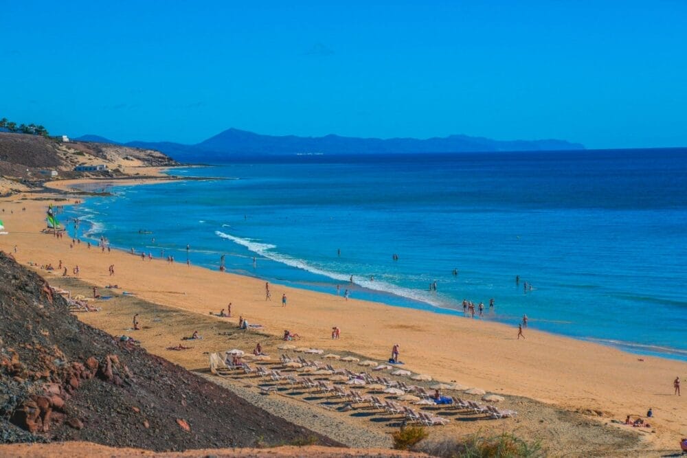 Un'ampia spiaggia sabbiosa con lettini e persone che nuotano, prendono il sole e passeggiano lungo la riva. Il mare limpido di Bravo Fuerteventura si estende fino all'orizzonte, con montagne lontane sotto un cielo azzurro brillante.