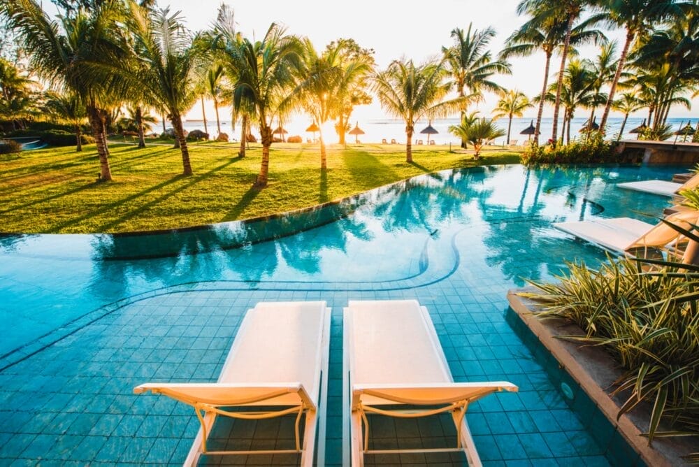 Due sedie a sdraio bianche si trovano accanto alla piscina turchese del Victoria Beachcomber Resort & Spa, circondate da palme e erba rigogliosa, con vista sulla spiaggia e sull'oceano al tramonto. La scena trasuda relax e lusso tropicale.