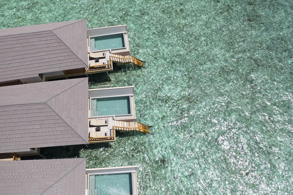 Vista aerea delle ville overwater con piscine private e ponti in legno del Brennia Kottefaru, un resort di lusso sulle isole Maldive, costruite sopra le limpide acque turchesi e collegate da scalinate che portano al mare.