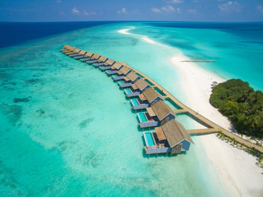 Vista aerea dei bungalow overwater sulla laguna turchese di Kuramathi Maldives, un'isola resort di lusso con spiaggia di sabbia bianca e vegetazione lussureggiante, circondata dall'oceano azzurro sotto un cielo soleggiato.