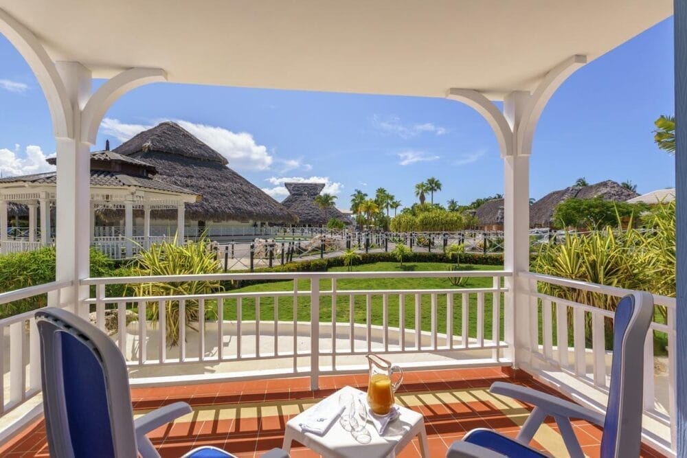 Il patio coperto del Royalton Hicacos Resort è caratterizzato da due sedie blu e da un tavolino con brocca e bicchieri, che si affacciano su un giardino lussureggiante con palme ed edifici dai tetti di paglia sotto un cielo blu brillante.