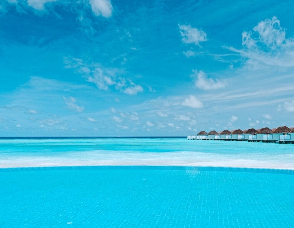 Una piscina a sfioro blu brillante si affaccia su un tranquillo oceano turchese al Nova Maldives. I bungalow overwater sono visibili sulla destra e il cielo è per lo più sereno con qualche nuvola sparsa.