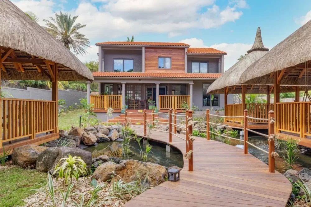 Una casa in stile tropicale con tetto di tegole rosse e accenti di legno, circondata da una vegetazione lussureggiante, evoca il fascino unico di un'esperienza di viaggio Zilwa Attitude, con passerelle di legno che conducono a gazebo con tetto di paglia su un tranquillo laghetto.