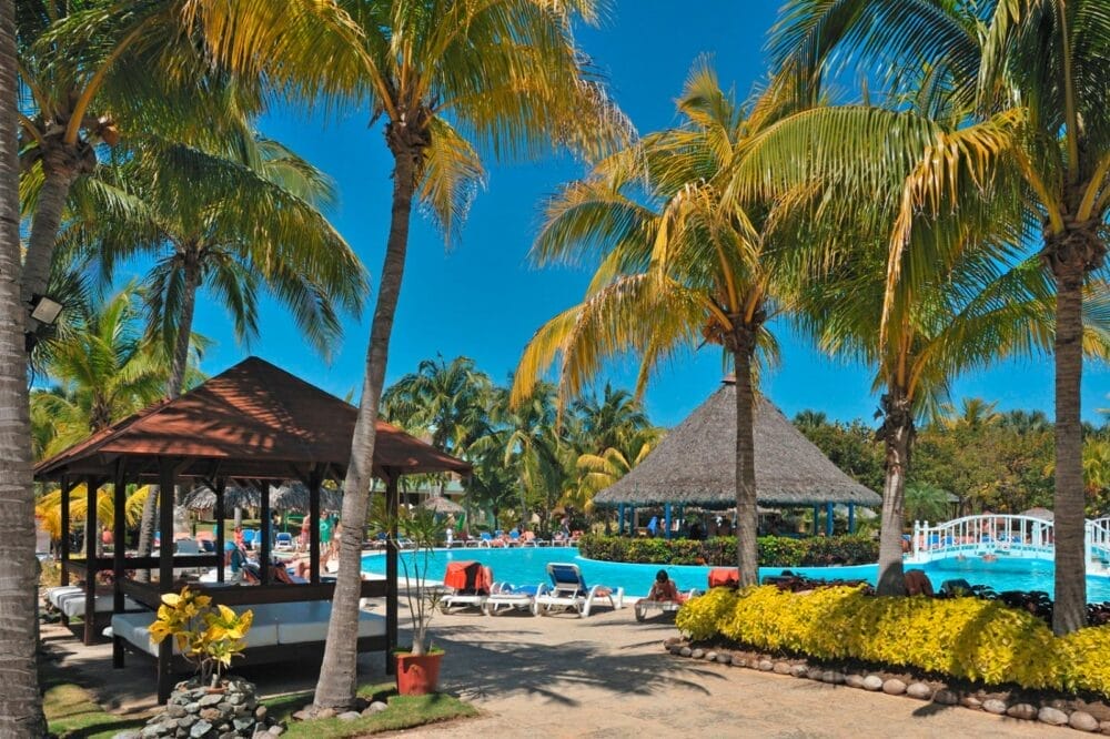 Un'area della piscina del resort tropicale Sol Palmeras con palme, lettini, un gazebo in legno e un bar con tetto in paglia. Le persone si rilassano a bordo piscina sotto un cielo azzurro e soleggiato, mentre cespugli e fiori costeggiano il camminamento.