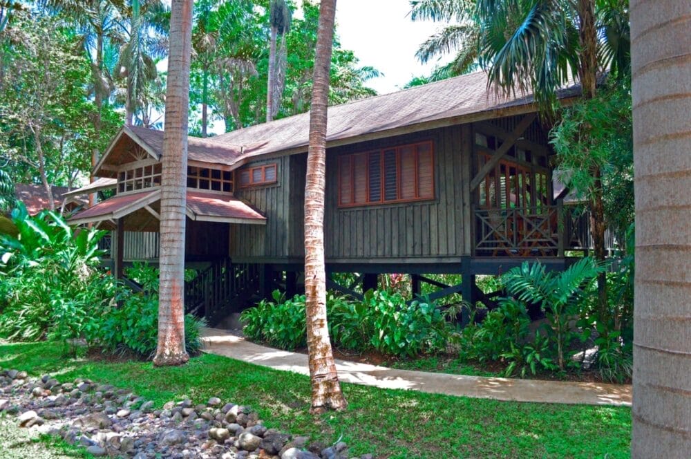 Una capanna di legno su palafitte circondata da piante tropicali verdi e alte palme, con un sentiero di pietra e un prato in primo piano, evoca l'atmosfera serena del Sunset At The Palms.