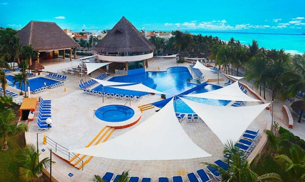 L'area della piscina del resort Wyndham Viva Maya è caratterizzata da vele ombreggianti bianche, sedie a sdraio blu, una vasca idromassaggio rotonda, palme tropicali, edifici con tetti di paglia e una vista mozzafiato sull'acqua turchese dell'oceano sotto un cielo blu brillante.