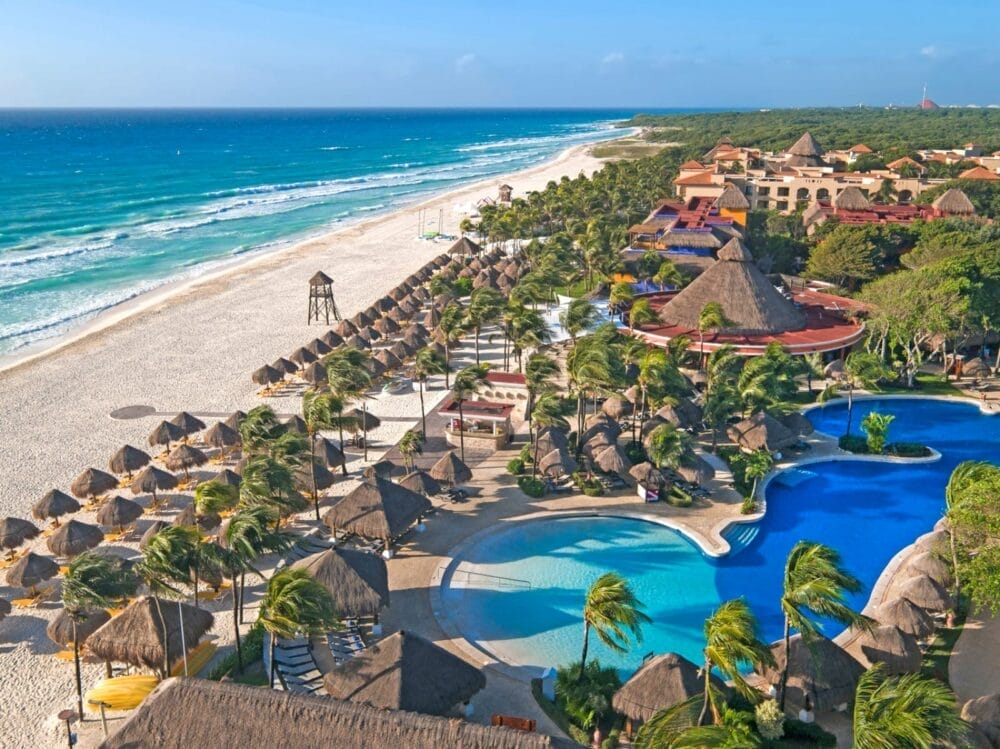 Vista aerea dell'Iberostar Waves Quetzal, un resort tropicale sulla spiaggia con edifici dai tetti di paglia, palme, diverse piscine e sedie a sdraio che costeggiano una spiaggia di sabbia bianca accanto alle onde turchesi dell'oceano sotto un cielo azzurro e limpido.