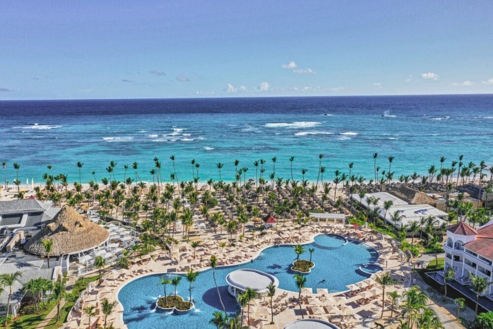 Vista aerea del resort Bahia Principe Luxury Ambar sulla spiaggia, caratterizzato da diverse piscine, sedie a sdraio, palme ed edifici con tetti di paglia, tutti affacciati su un oceano turchese sotto un cielo azzurro e limpido.