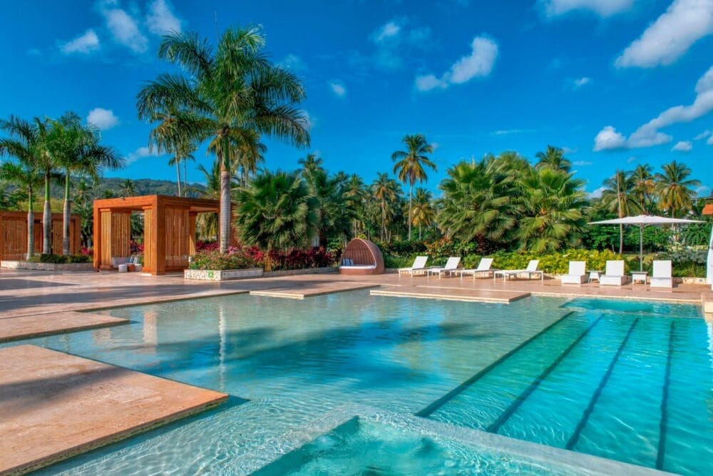 La lussuosa piscina all'aperto del Viva V Samana by Wyndham, circondata da palme, sedie a sdraio, cabanas e cielo azzurro, crea un'atmosfera tropicale e rilassante.