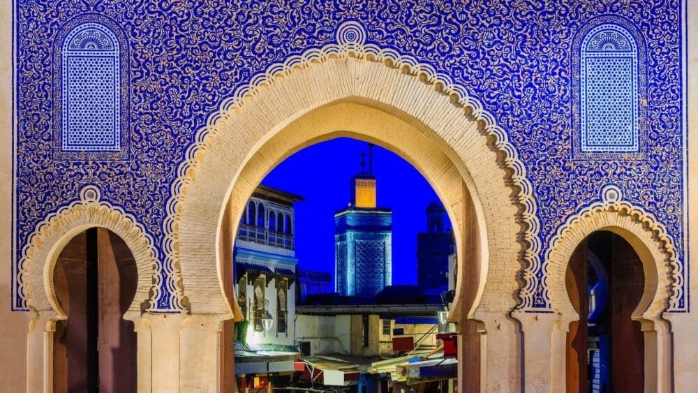 Cancello ornato di piastrelle blu con intricati motivi arabi, grandi arcate e una vista di moschee ed edifici illuminati di notte - una scena incantevole spesso presente in un Gran Tour Marocco attraverso il cuore storico di Marrakech.