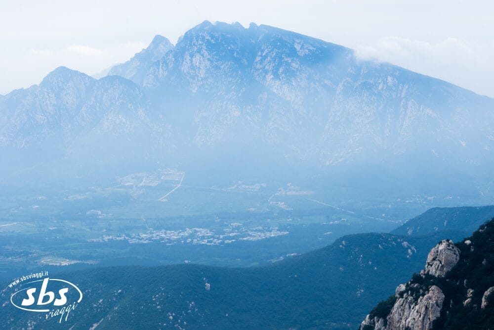 Una vista nebbiosa di una grande e aspra catena montuosa che sovrasta una valle verde, con una piccola città sottostante. Perfetta per un Tour Cina storica, la scena è parzialmente oscurata dalla foschia. Il logo "sbs viaggi" è in basso a sinistra.