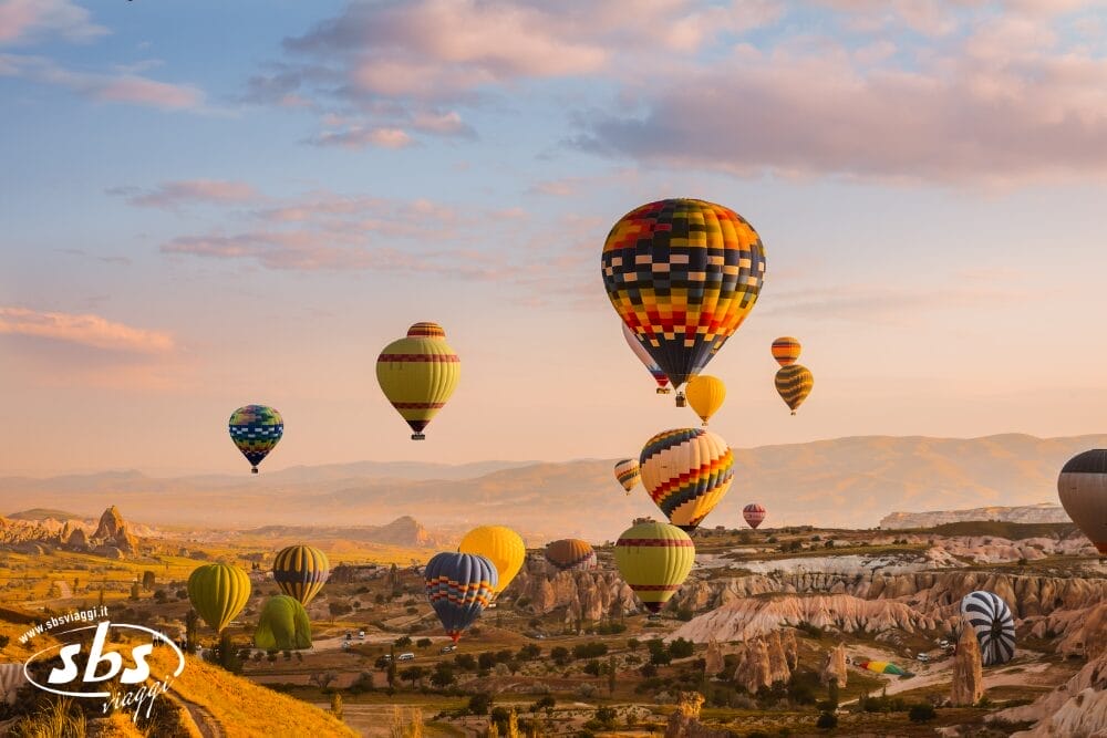 Colorate mongolfiere fluttuano sopra uno scenografico paesaggio roccioso all'alba, con nuvole soffici e colline lontane. Questa vista pacifica e vibrante cattura la bellezza del volo in mongolfiera durante il Tour Turchia attraverso la Turchia Iconica.
