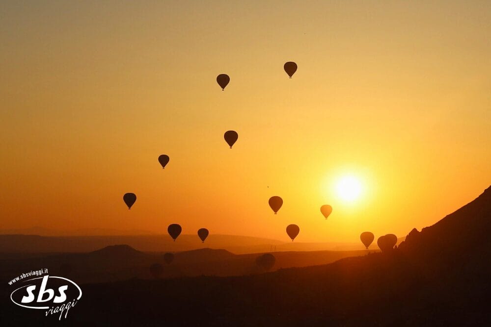 Le mongolfiere fluttuano nel cielo all'alba sopra la Cappadocia, stagliandosi contro un orizzonte giallo-arancio. Le colline ondulate si estendono sotto di noi, invitandovi a partecipare a un tour del 2026. Il logo di SBS Viaggi appare nell'angolo.