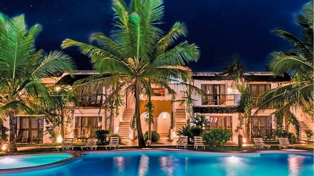 Un resort tropicale di notte, il My Blue Hotel è caratterizzato da palme, una grande piscina illuminata e un edificio a due piani con balconi luminosi, il tutto circondato da una vegetazione lussureggiante.