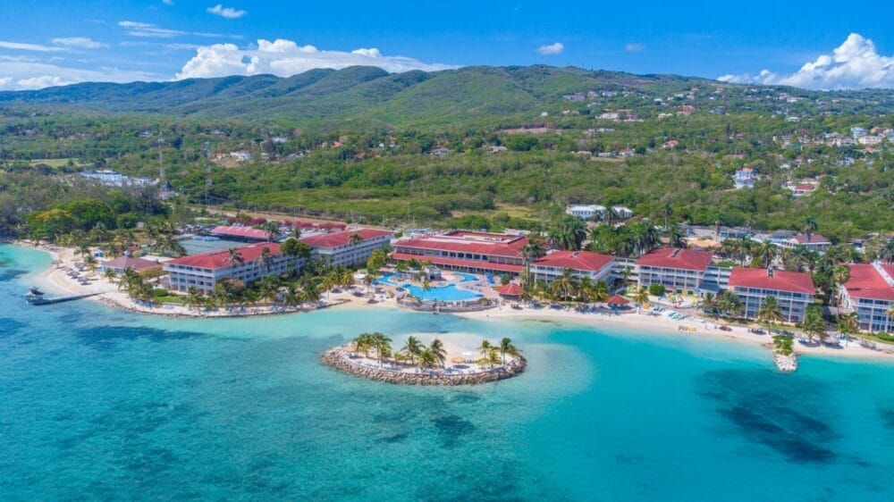 Veduta aerea del Catalonia Montego Bay, un resort tropicale sulla spiaggia con edifici dai tetti rossi, piscine, palme, spiagge sabbiose e acque turchesi, immerso in colline verdi sotto un cielo blu brillante.