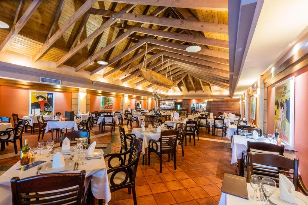 Un ristorante spazioso in un resort sulla spiaggia di Punta Cana, con travi in legno sul soffitto, tavoli ordinati, sedie in legno e opere d'arte colorate alle pareti. Le luci calde creano un'atmosfera accogliente, perfetta dopo una giornata di relax alle terme.
