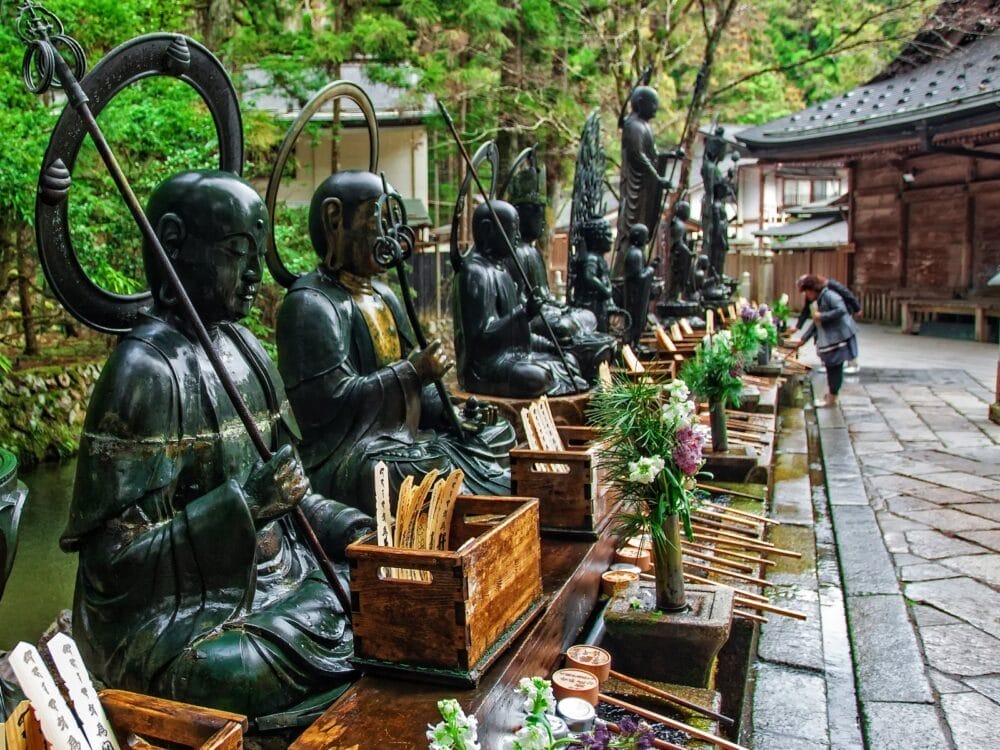 Una fila di statue scure, probabilmente monaci buddisti, siedono all'aperto con cassette di legno per le offerte e fiori davanti. Si vede una persona che fa un'offerta o rende omaggio. Il verde lussureggiante e gli edifici tradizionali circondano la scena.