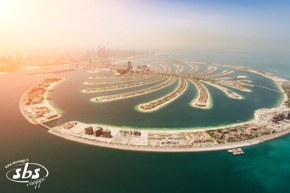 Vista aerea di Palm Jumeirah, un'isola artificiale a forma di palma a Dubai, con i grattacieli sullo sfondo e l'acqua turchese che circonda l'isola. Il logo di SBS Viaggi appare nell'angolo in basso a sinistra, evidenziando il vostro prossimo tour a Dubai.