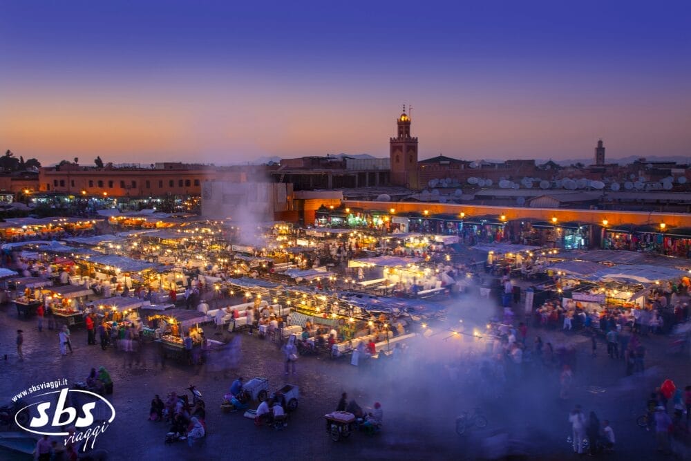 Un vivace mercato notturno a Marrakech, in Marocco, con bancarelle illuminate, fumo che sale dai venditori di cibo e persone che esplorano queste meraviglie. La torre di una moschea e gli edifici della città sono visibili sullo sfondo al tramonto.