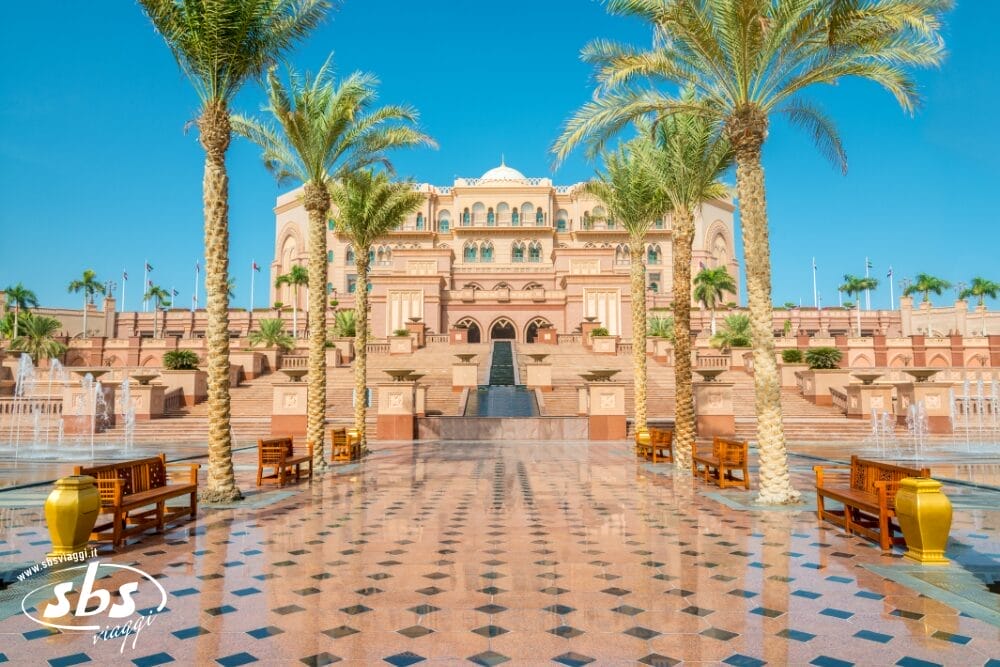 Un grande edificio decorato con finestre ad arco e balconi si erge dietro palme, fontane e camminamenti di piastrelle decorate sotto un cielo azzurro e limpido: una scena perfetta per il turismo emiratino o per uno splendido Grand Tour emiratino. Le panchine costeggiano il percorso che porta all'ingresso.