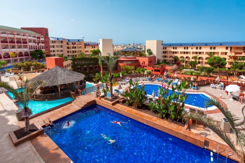 Vista aerea del resort Alpiclub Jacaranda con diverse piscine, palme, lettini, un bar con tetto di paglia e persone che nuotano e si rilassano sotto un cielo azzurro.
