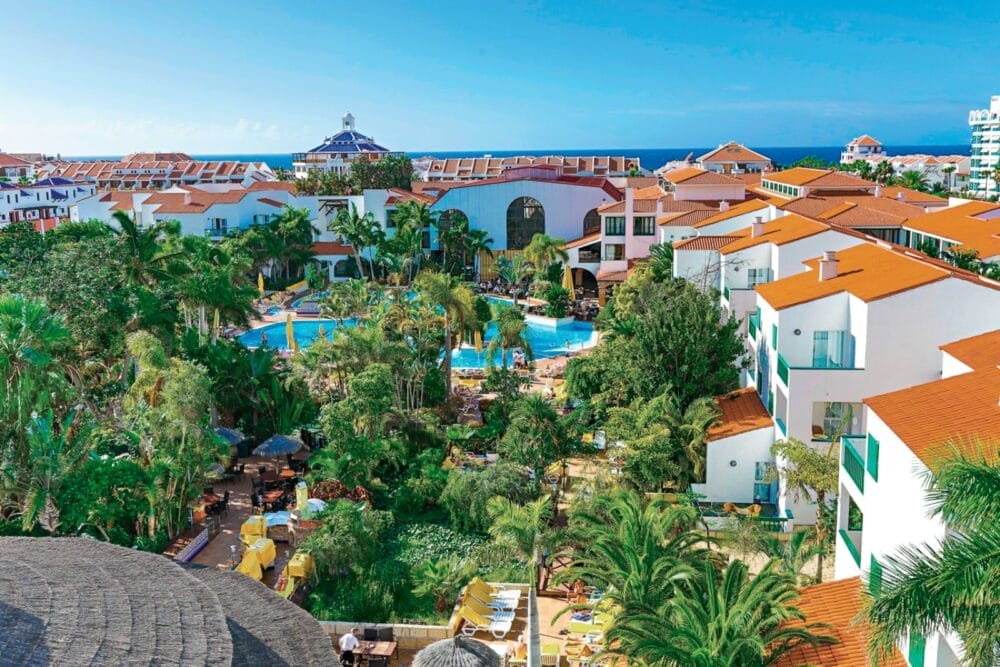 Vista aerea del Park Club Europe, un resort Alpiclub con edifici dai tetti rossi, palme, giardini lussureggianti e una grande piscina blu, il tutto immerso in un cielo limpido vicino all'oceano.