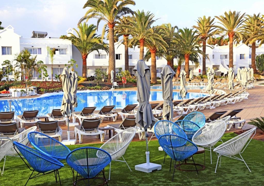 L'area della piscina dell'Alpiclub Corralejo è caratterizzata da file di sedie a sdraio e ombrelloni chiusi, circondati da palme. Moderne sedie bianche e blu si trovano sull'erba in primo piano, con edifici bianchi sullo sfondo.