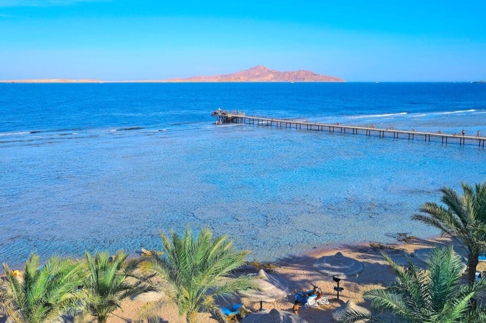 Un paradiso tropicale vi attende al Bravo Tamra Beach, dove l'acqua cristallina, le palme e gli ombrelloni di paglia costeggiano la riva. Un lungo molo di legno conduce al mare, con una montagna lontana visibile sotto il cielo azzurro.