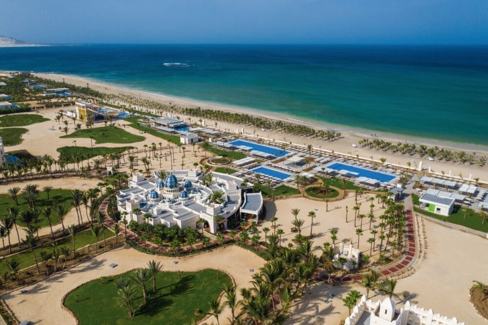 Vista aerea del Searesort Riu Karamboa, un lussuoso resort sulla spiaggia con un grande edificio bianco, palme, prati verdi, aree piscina e lettini che costeggiano un litorale sabbioso accanto alle acque turchesi dell'oceano sotto un cielo azzurro e limpido.