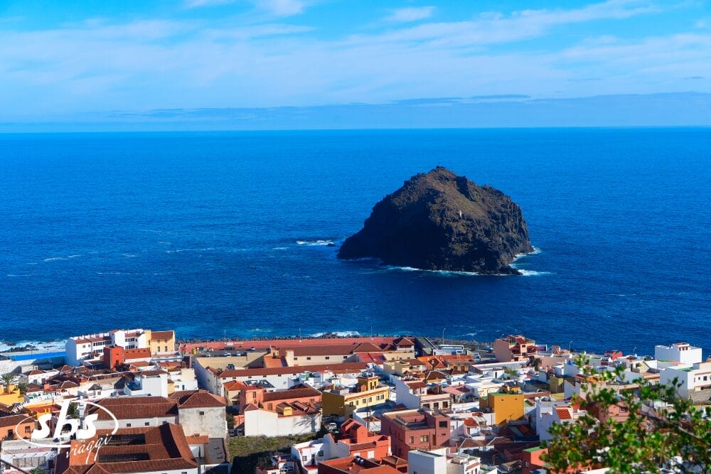Colorata cittadina costiera con edifici dai tetti rossi che si affacciano sul blu intenso dell'oceano e su un'isola rocciosa, perfetta per vivere le Meraviglie di Tenerife durante uno Speciale Capodanno sotto un cielo limpido e luminoso.