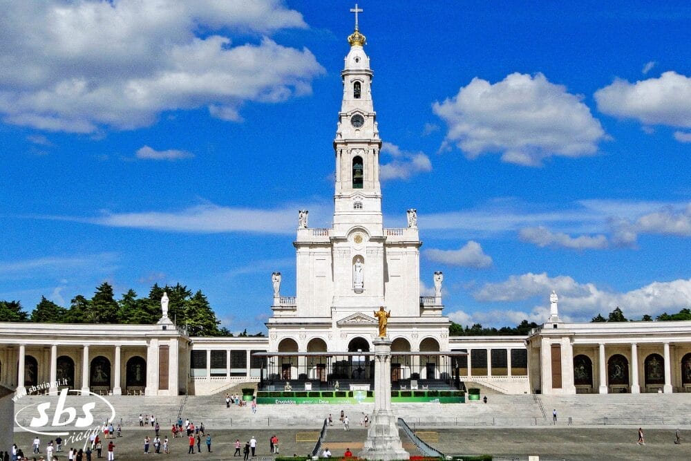 L'immagine mostra il Santuario di Nostra Signora di Fátima a Portogallo, con la sua imponente basilica bianca, l'alto campanile centrale sormontato da una croce e le persone che passeggiano nell'ampia piazza sotto un cielo azzurro con nuvole sparse.