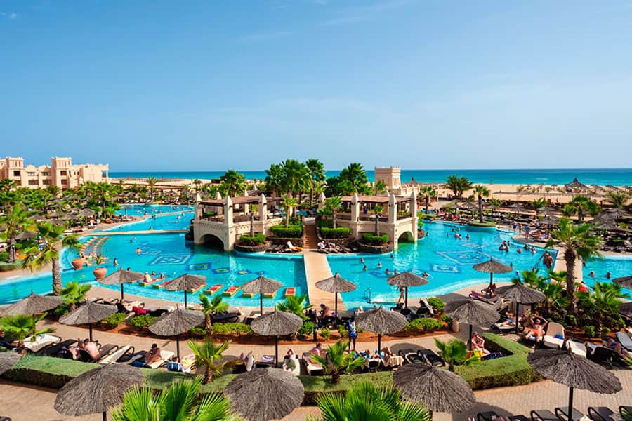 Una grande piscina del resort con palme, lettini e ombrelloni di paglia, circondata da persone che nuotano e si rilassano. Gli edifici del resort e l'oceano sono visibili sullo sfondo sotto un cielo azzurro e limpido.