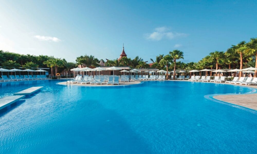 La grande piscina blu chiaro del Riu Cabo Verde è circondata da palme, lettini e ombrelloni bianchi sotto il cielo luminoso di questo resort tropicale.