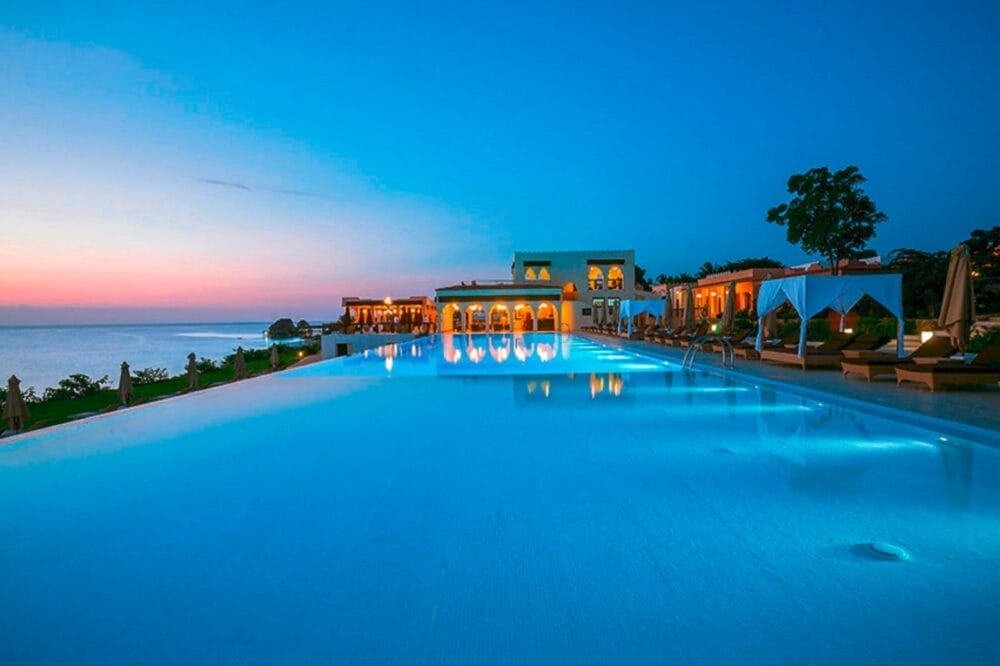 Piscina a sfioro al tramonto con luci blu scintillanti, sedie a sdraio e cabine. Gli eleganti edifici del resort Riu Palace Zanzibar sono illuminati e si affacciano sul mare calmo e sul cielo colorato della sera.