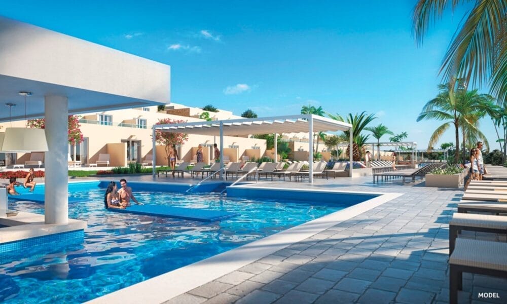 La moderna piscina all'aperto del Riu Jambo Hotel è dotata di sedie a sdraio, ombrelloni, palme e un bar a bordo piscina. Una coppia si rilassa in piscina sotto un cielo azzurro, creando un'atmosfera da resort di lusso.
