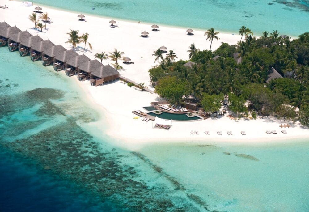 Veduta aerea del Constance Moofushi, un resort di lusso alle Maldive, con bungalow overwater, spiaggia di sabbia bianca, acqua limpida e turchese, lettini, palme e una vivace barriera corallina.