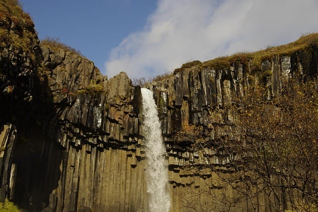 Un'alta cascata scende da scure colonne di basalto, circondata da un rado fogliame autunnale, sotto un cielo azzurro parzialmente nuvoloso: una scena stupefacente che sembra quasi una bozza automatica dell'arte della natura.