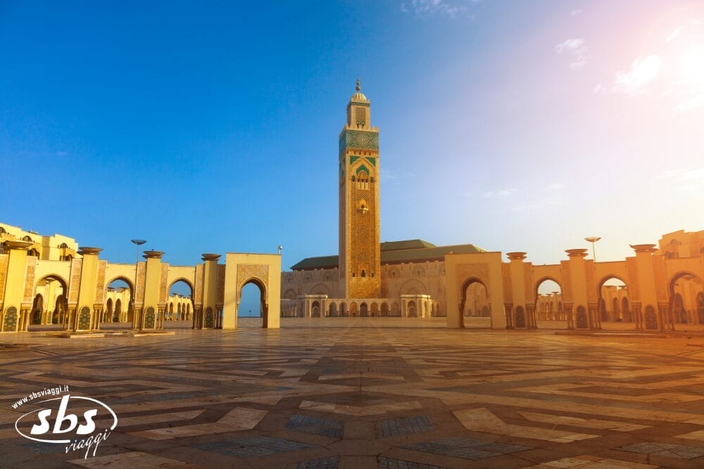 Una vista grandangolare della Moschea di Hassan II a Casablanca, in Marocco, con il suo alto minareto, gli archi ornati e il cortile decorato, sotto un cielo azzurro e limpido all'alba: una tappa indimenticabile di qualsiasi tour VIP.