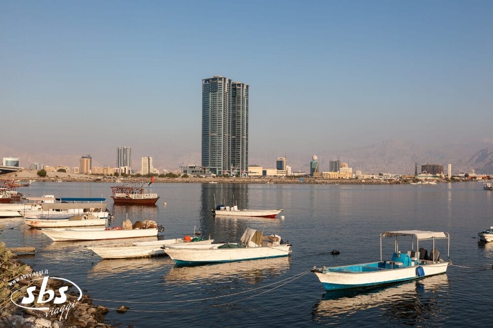 Diverse piccole imbarcazioni galleggiano sull'acqua calma di un porto, con lo skyline della città e un alto grattacielo moderno sullo sfondo, sotto un cielo limpido: una vista iconica del minitour Mare Ras Al Khaimah. Il logo "sbs" appare nell'angolo in basso a sinistra.