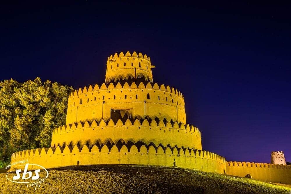 Forte rotondo a più livelli con merli appuntiti, illuminato da luci gialle contro un cielo notturno blu scuro: una vista iconica per ogni Grand Tour emiratino. Gli alberi si allineano sulla sinistra, mentre una torre più piccola si erge in lontananza sulla destra.