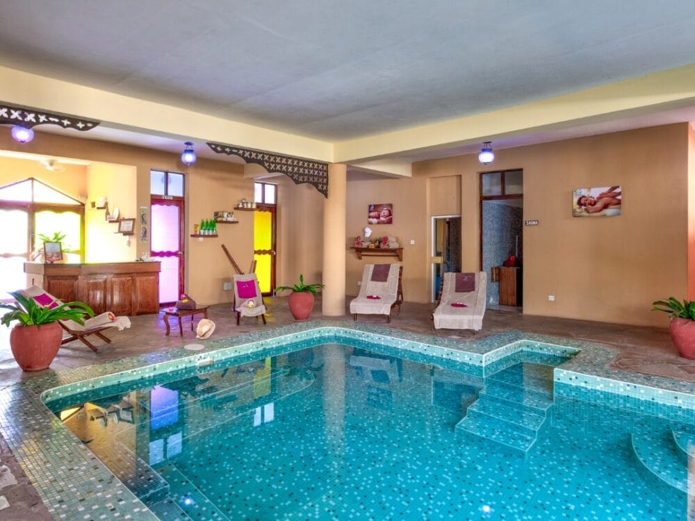 Area spa interna del Waridi Beach Resort con piccola piscina a mosaico, gradini incorporati, sedie a sdraio, piante in vaso e pareti di colore giallo e marrone caldo. Le luci soffuse e le vetrate colorate creano un'atmosfera rilassante da spa.