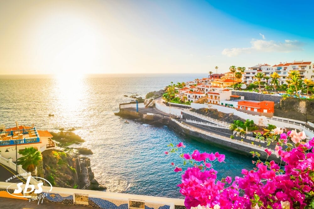 Una colorata cittadina costiera di Tenerife con edifici bianchi e arancioni che si affacciano sull'oceano al tramonto, fiori rosa vibranti in primo piano e la luce del sole che si riflette sull'acqua: un luogo perfetto per un tour di Capodanno.