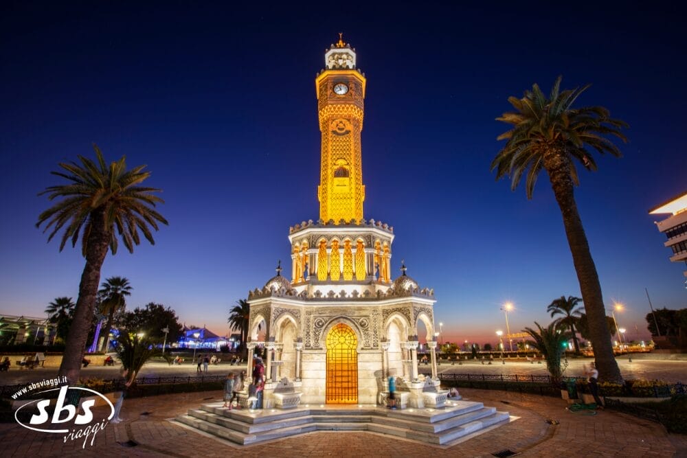 La Torre dell'Orologio di Smirne, illuminata, si erge in una piazza pubblica all'imbrunire, circondata da palme, con un cielo blu intenso e le luci della città sullo sfondo: una tappa obbligata del vostro viaggio Grantour Turchia 2026.