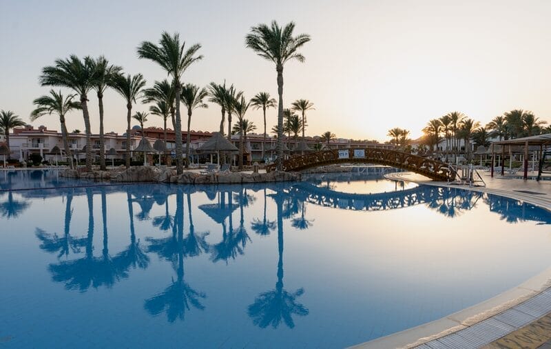 All'alba, la piscina del Parrotel Beach Resort brilla con i riflessi delle palme, una passerella in legno e i lettini, sullo sfondo dello splendido skyline di Sharm El Sheikh e dei giardini lussureggianti.