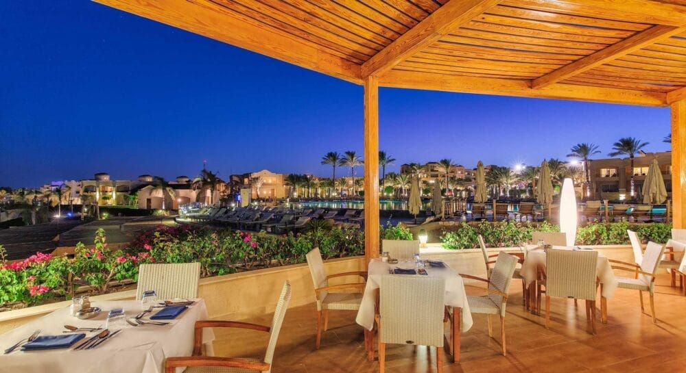 Il patio del ristorante all'aperto del Cleopatra Luxury Resort è caratterizzato da tavoli e sedie bianche che si affacciano sulla piscina del resort, sulle palme e sugli edifici al tramonto, con luci soffuse e fiori vivaci lungo la terrazza sotto un cielo blu intenso.
