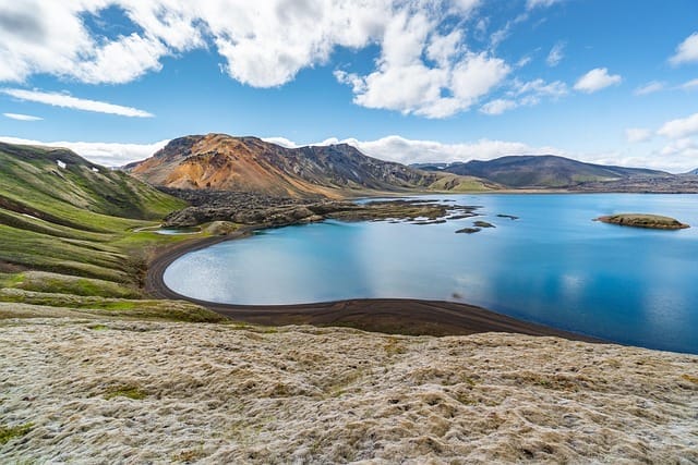 Un sereno lago azzurro circondato da verdi colline e aspre montagne multicolori, sotto un cielo parzialmente nuvoloso, con l'acqua calma che riflette il paesaggio: una cornice perfetta per un momento di Bozza automatica.