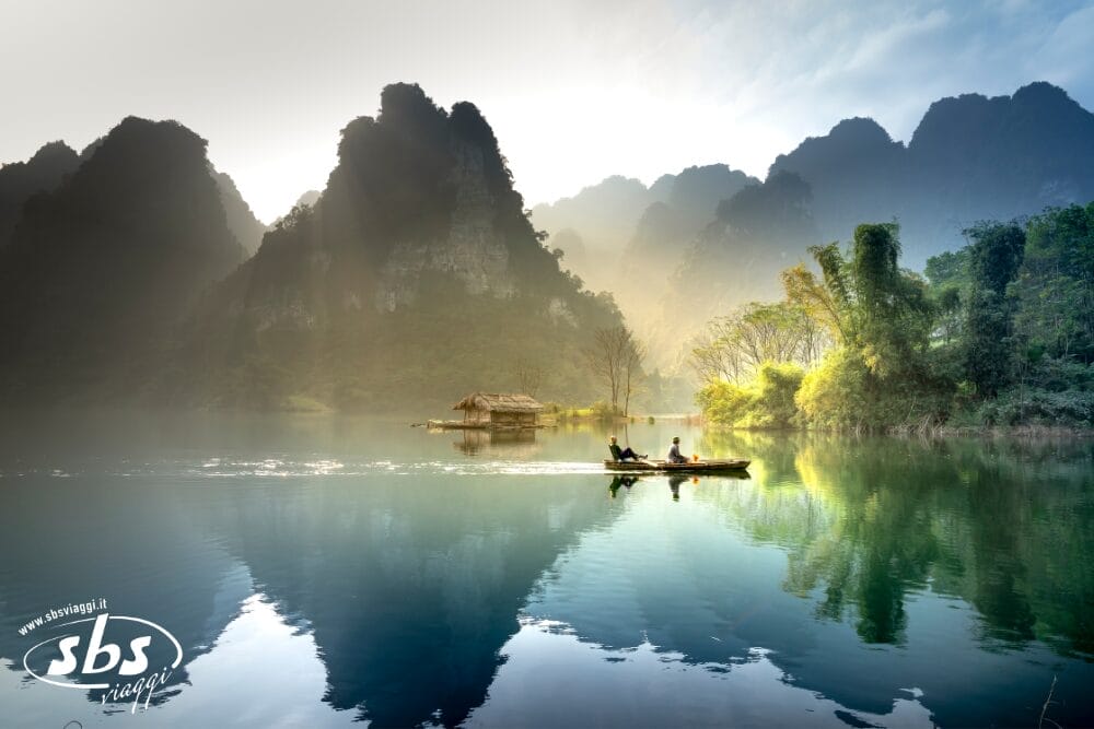 Due persone remano in canoa su un lago calmo e riflessivo, circondato da montagne nebbiose e da una vegetazione lussureggiante, mentre una piccola capanna di legno riposa sulla riva sotto una luce solare morbida e dorata, immergendo la scena in un'atmosfera tranquilla e bozza automatica.