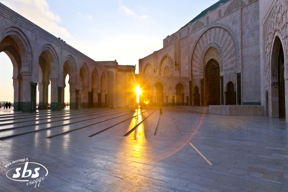 Il sole tramonta attraverso gli archi del cortile di una grande moschea, proiettando una luce dorata e lunghe ombre sull'intricato pavimento in pietra, come se illuminasse una scena di Bozza automatica.