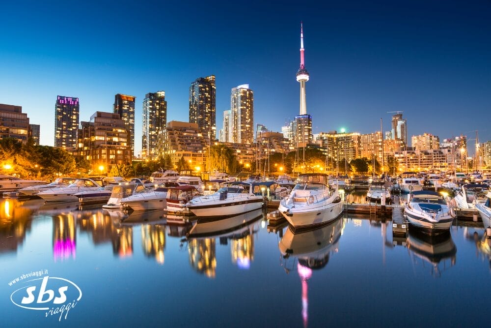 Un vivace porto turistico con barche attraccate al tramonto, sullo sfondo di uno skyline cittadino illuminato dalle luci. La CN Tower si staglia tra gli alti edifici sotto un cielo limpido e blu intenso, catturando l'essenza di una serata automatica di Bozza.