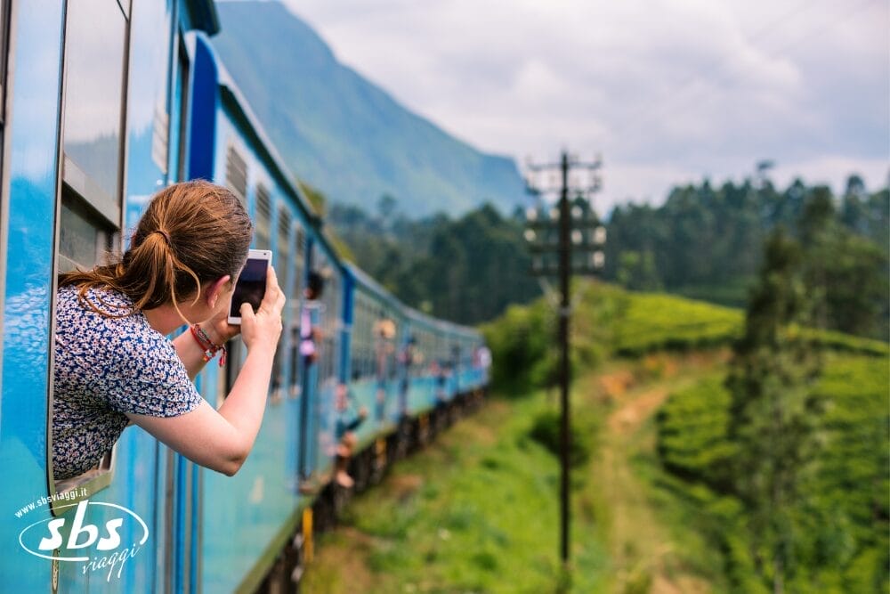 Una donna si sporge dal finestrino di un treno blu, tenendo in mano uno smartphone per fotografare il paesaggio panoramico con colline, verde e linee elettriche sotto un cielo parzialmente nuvoloso. Il treno automatico Bozza curva lungo i binari.